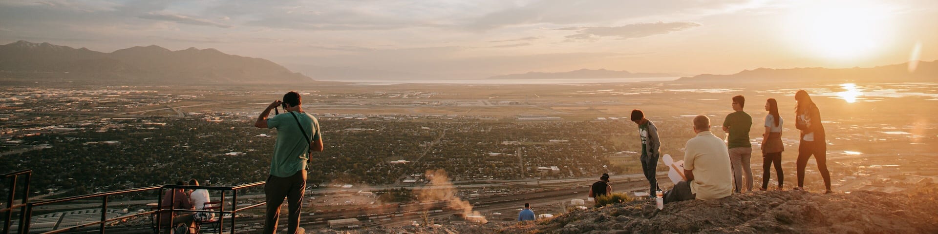 A group of people on a hill overlooking a cityscape with mountains in the distance during sunset.