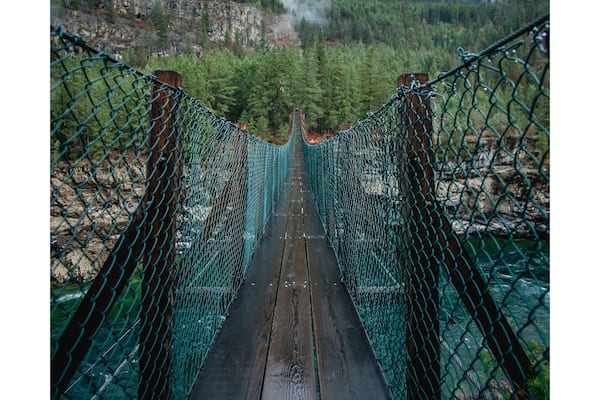 A suspension bridge with a wooden walkway, surrounded by forested hills and a rocky cliff.