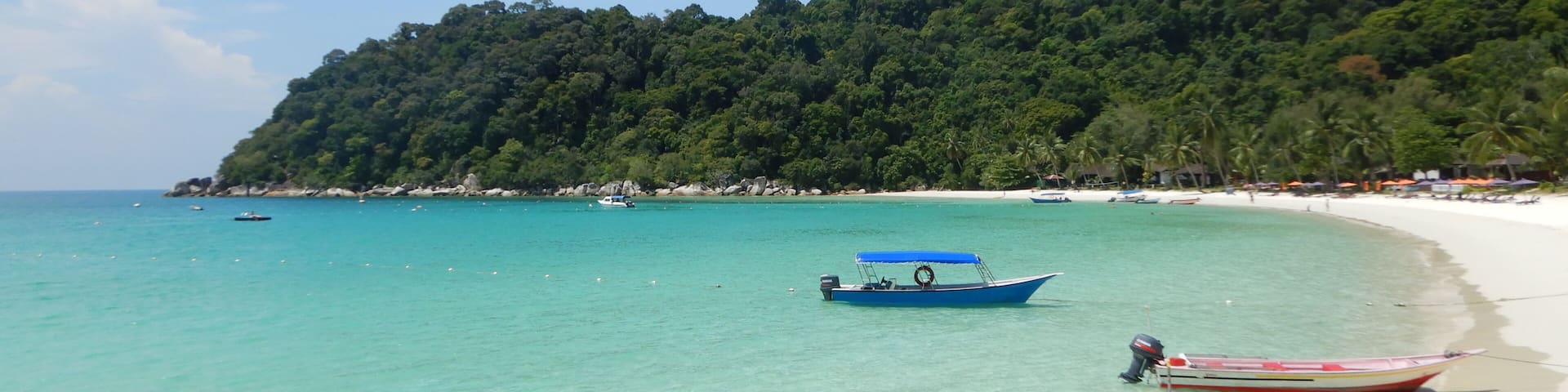 A beach with clear turquoise water, a sandy shore, and a forested hill in the background.