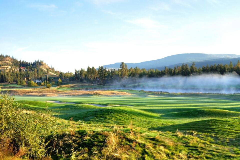 A golf course with rolling green hills, a forested area, and distant mountains.