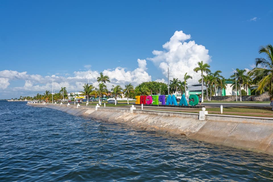 A coastal scene with a calm sea, palm trees, and colorful buildings along the shore.