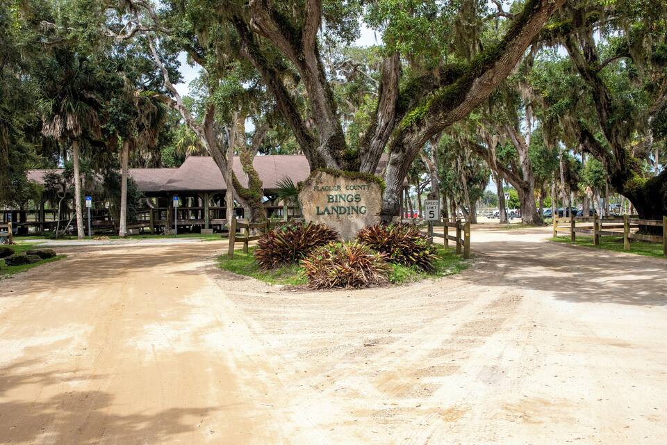 A dirt road leading to a sign reading "Flagler County Bings Landing" surrounded by trees and a building.