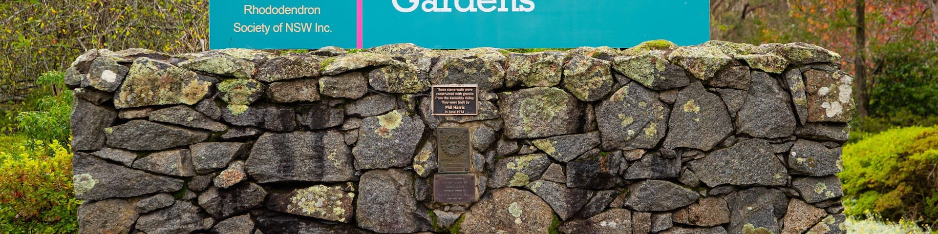 A sign for Campbell Rhododendron Gardens with a stone wall in the foreground.