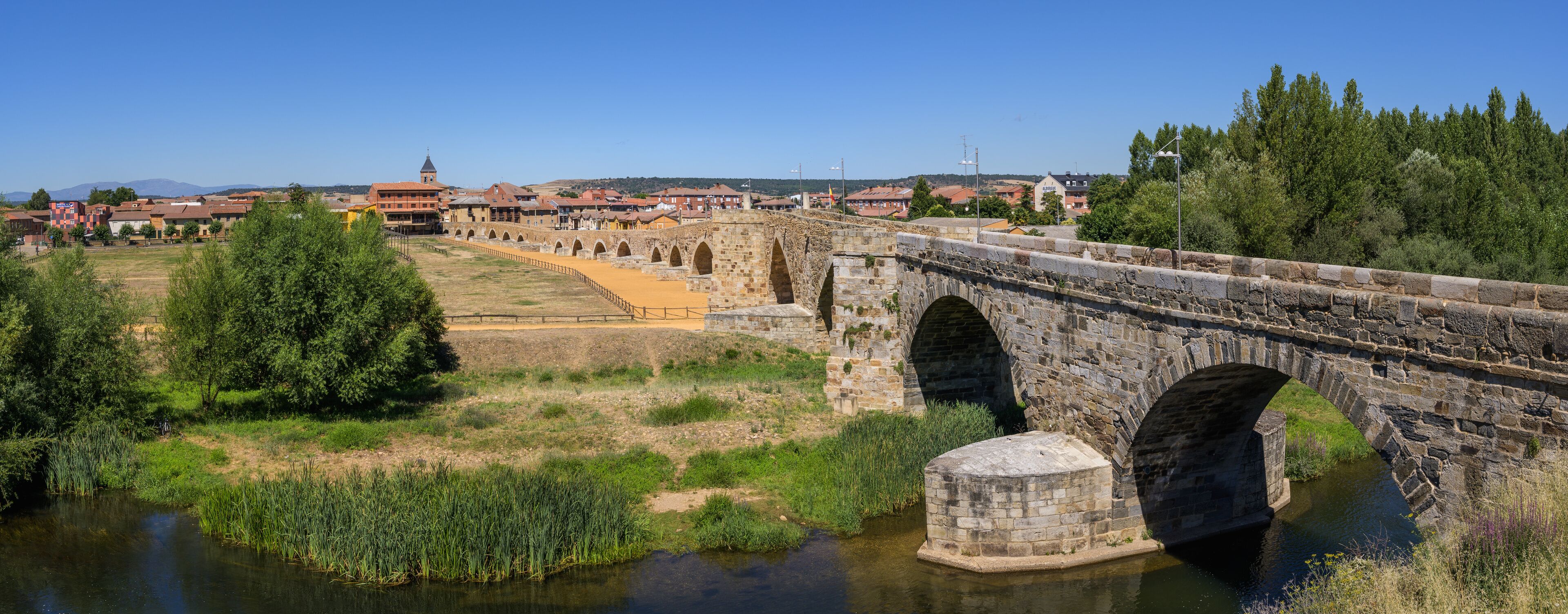 A stone bridge over a river, surrounded by greenery and a town in the background.