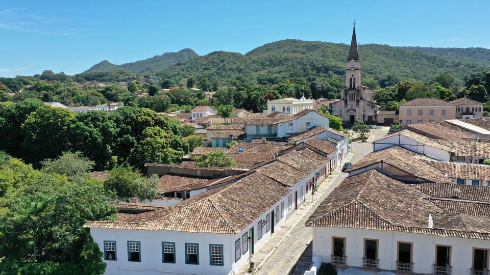 A town with terracotta-roofed buildings, a church with a tall steeple, and lush green hills in the background.