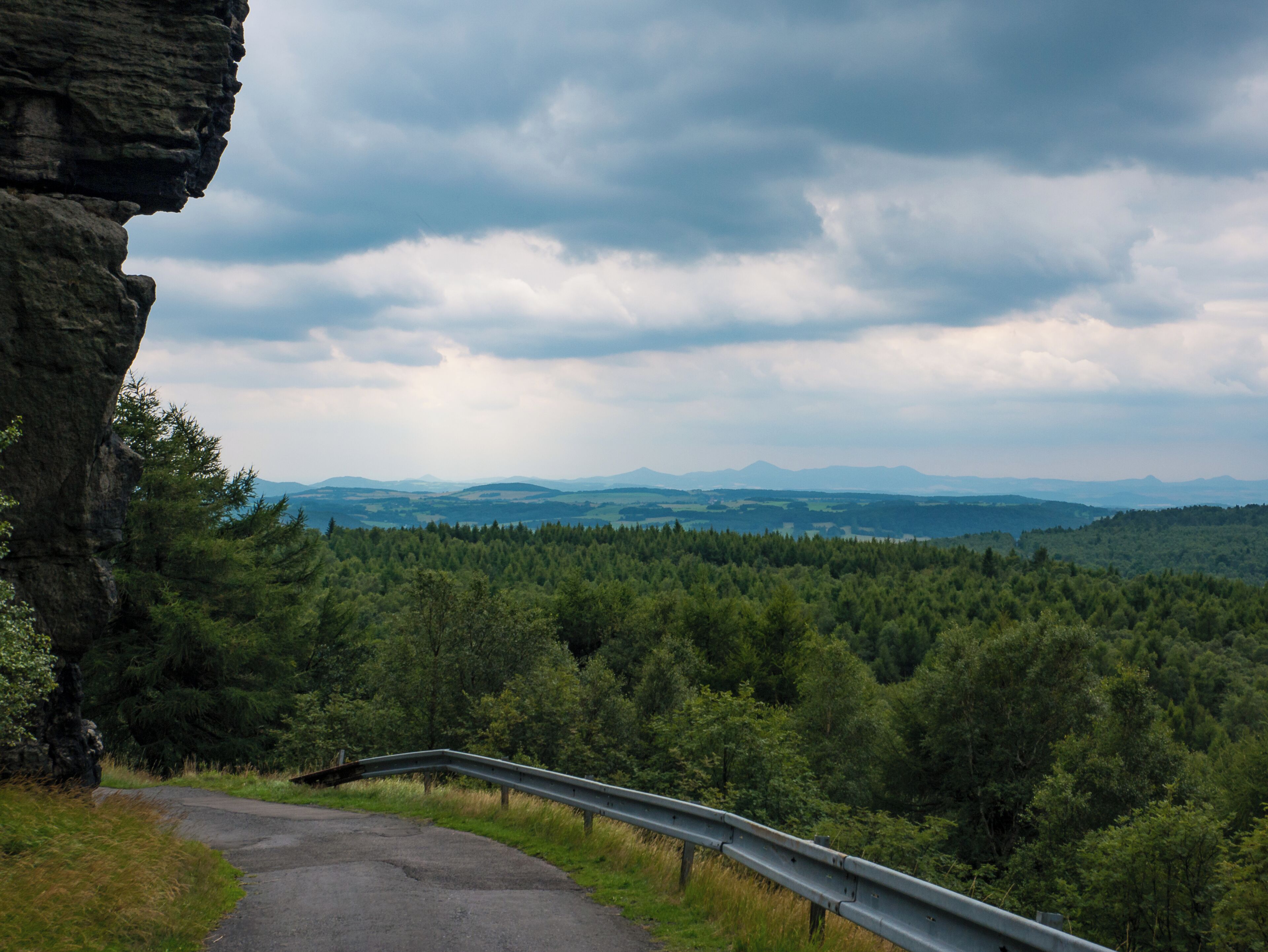 A winding road with a guardrail, surrounded by dense forest and mountains in the distance under a cloudy sky.