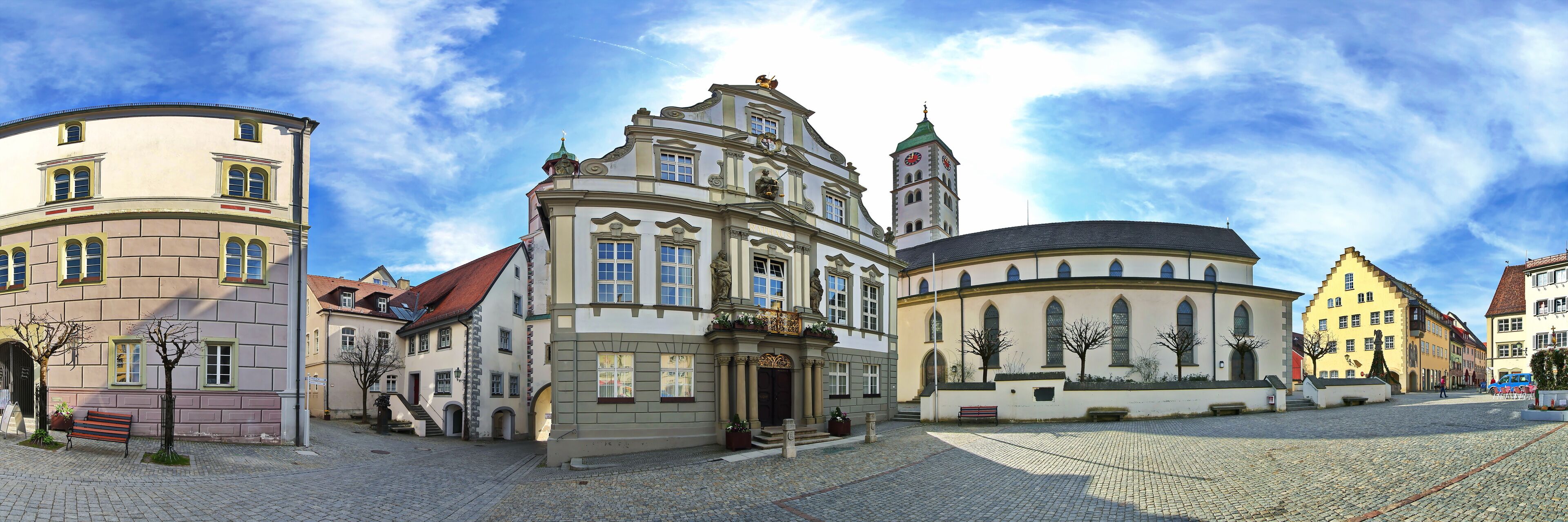 A historic European town square with cobblestone streets, traditional buildings, and a prominent church with a clock tower.