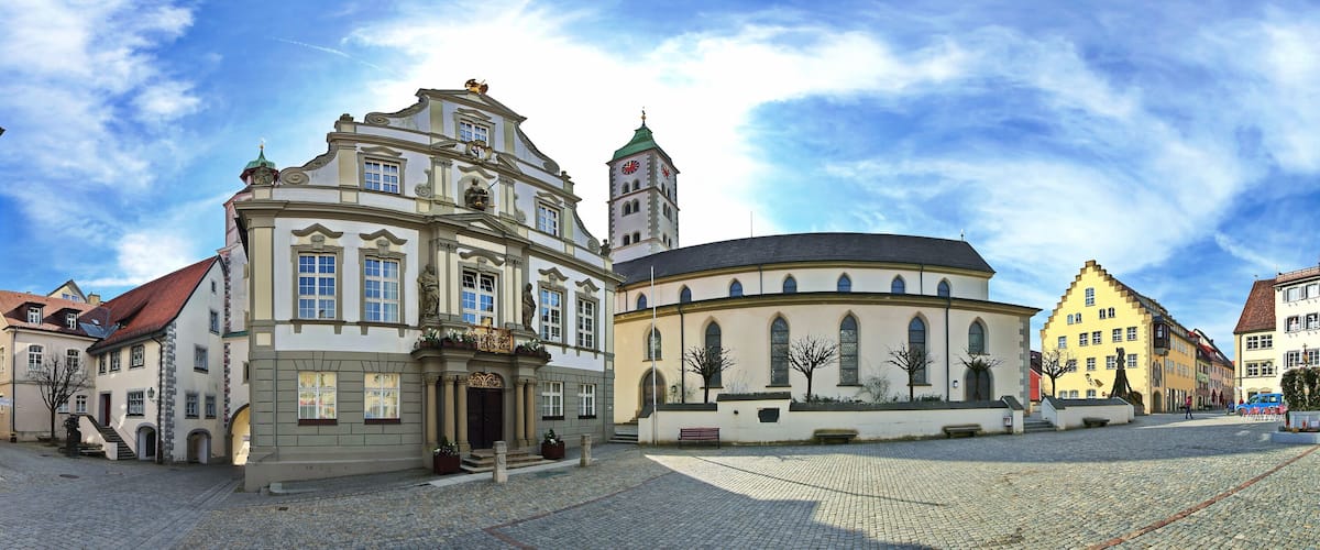 A historic European town square with cobblestone streets, traditional buildings, and a prominent church with a clock tower.