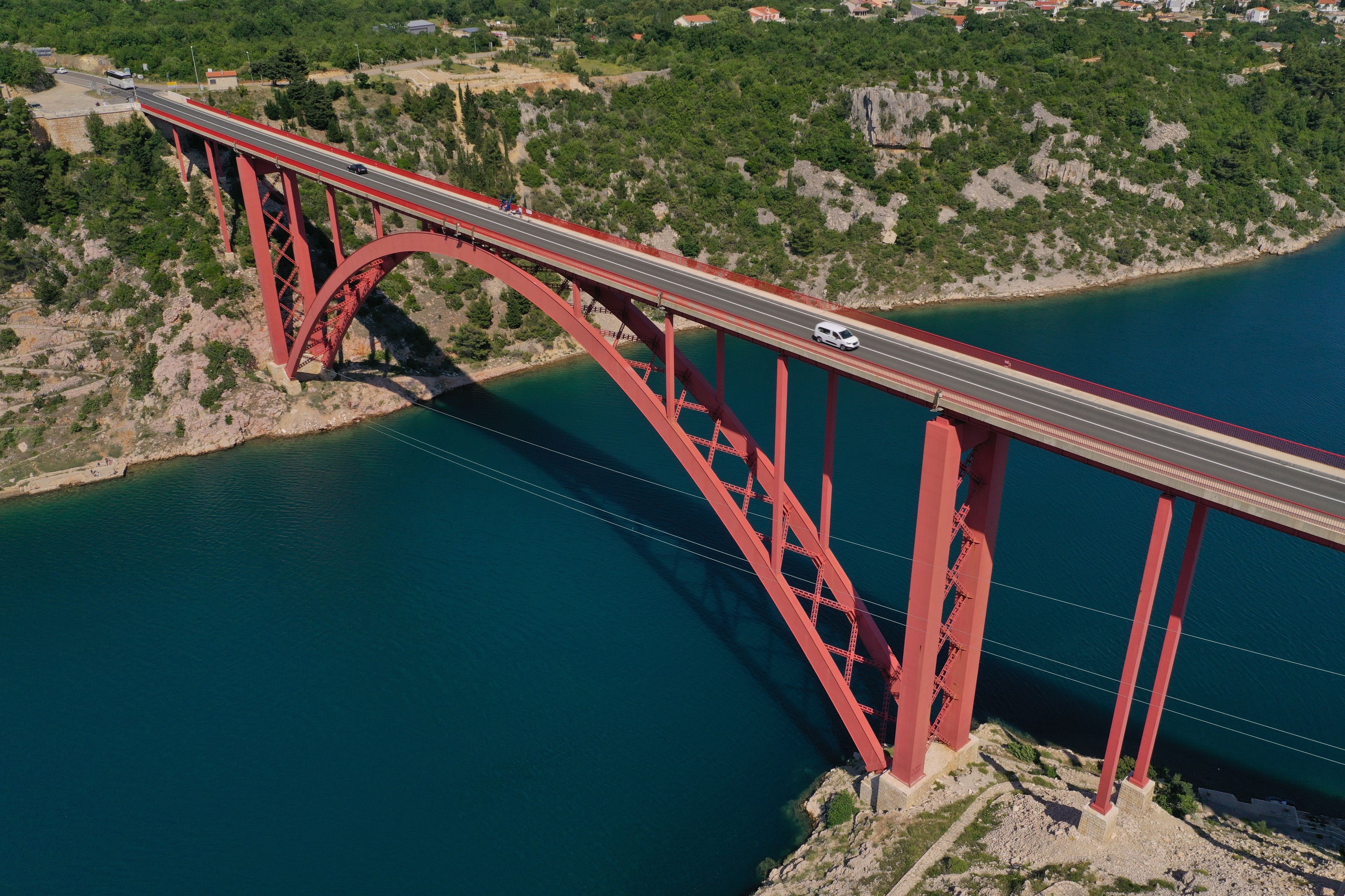 Aerial view of the red Maslenica bridge spanning across the deep blue water of the Maslenica Strait with rocky shores and green hills in Jasenice, Croatia.