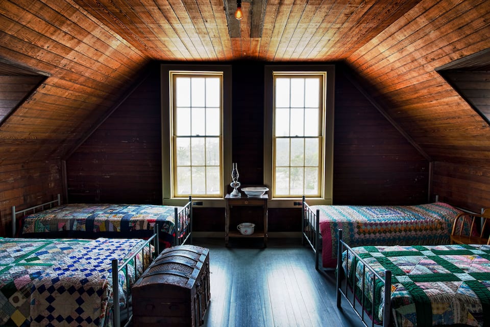 A rustic attic bedroom with wooden walls and sloped ceiling, two windows, and three beds with quilts.
