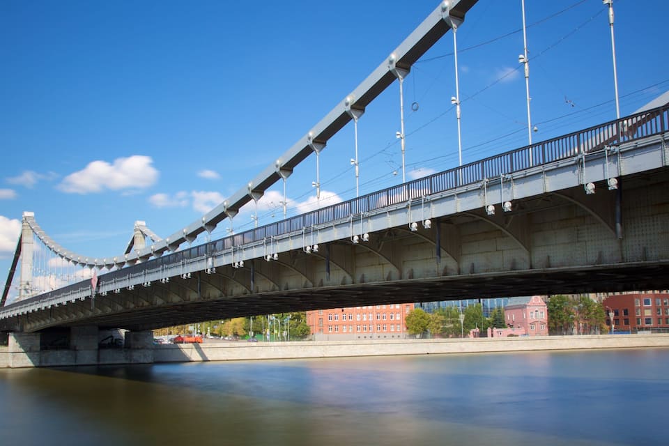 A large suspension bridge spanning a calm river, with a cityscape in the background.