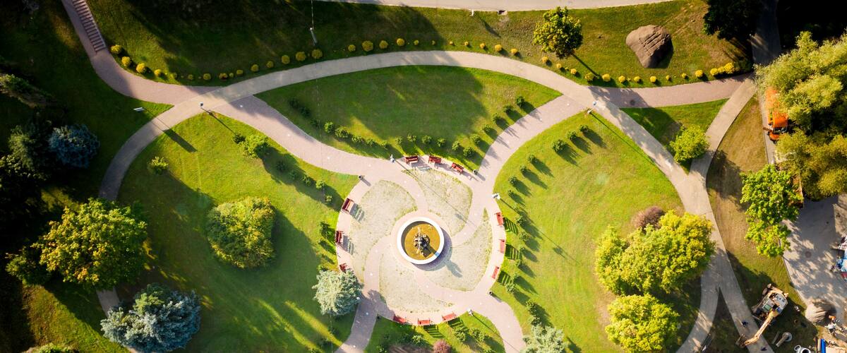 An aerial view of a park with a circular fountain, pathways, and surrounding greenery.