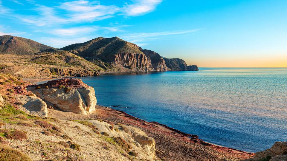 A coastal landscape with a rocky beach, a clear blue sea, and a mountain in the background.