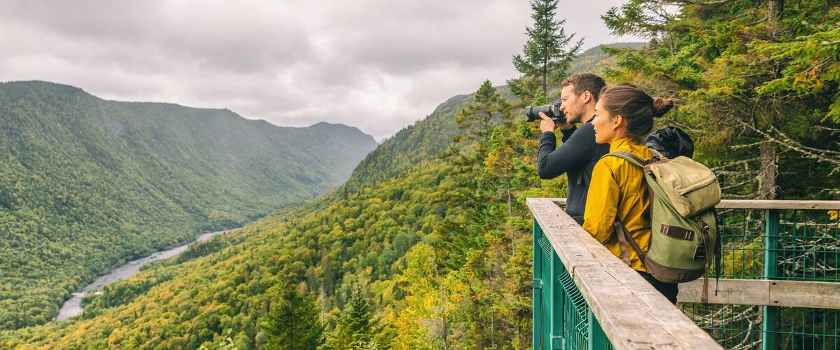 Travel couple hikers tourists taking photo with camera at view of mountain landscape in Autumn forest Parc de la Jacques Cartier, Quebec, Canada. Panorama banner background.