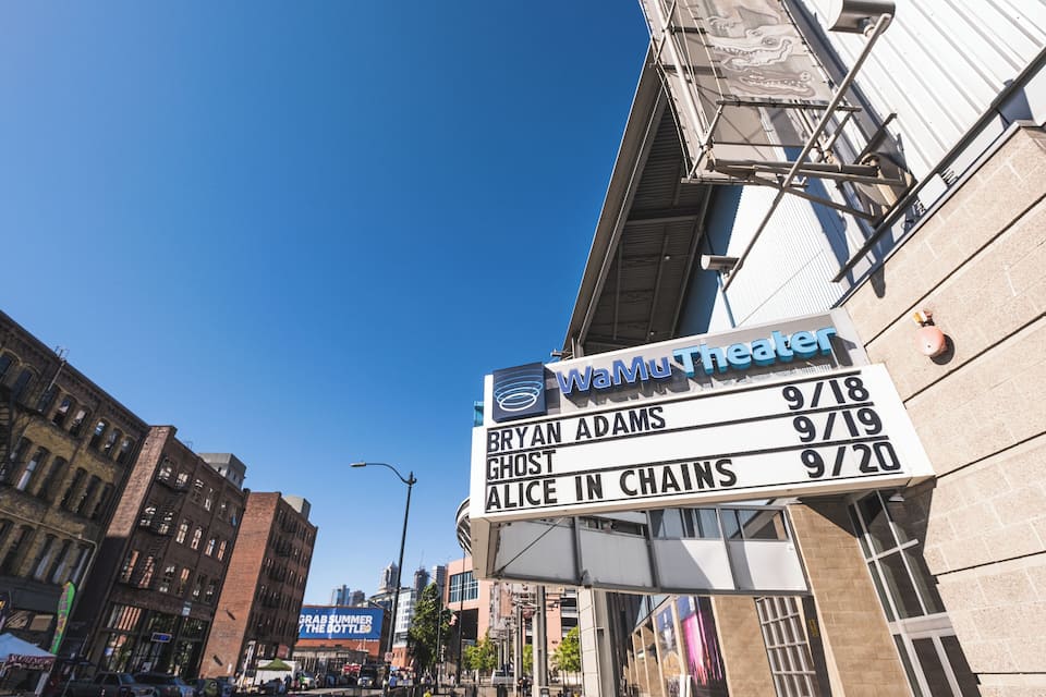 Un cartel de cine con el nombre "WaMu Theater" y una lista de espectáculos próximos, incluyendo "Bryan Adams", "Ghost" y "Alice in Chains".
