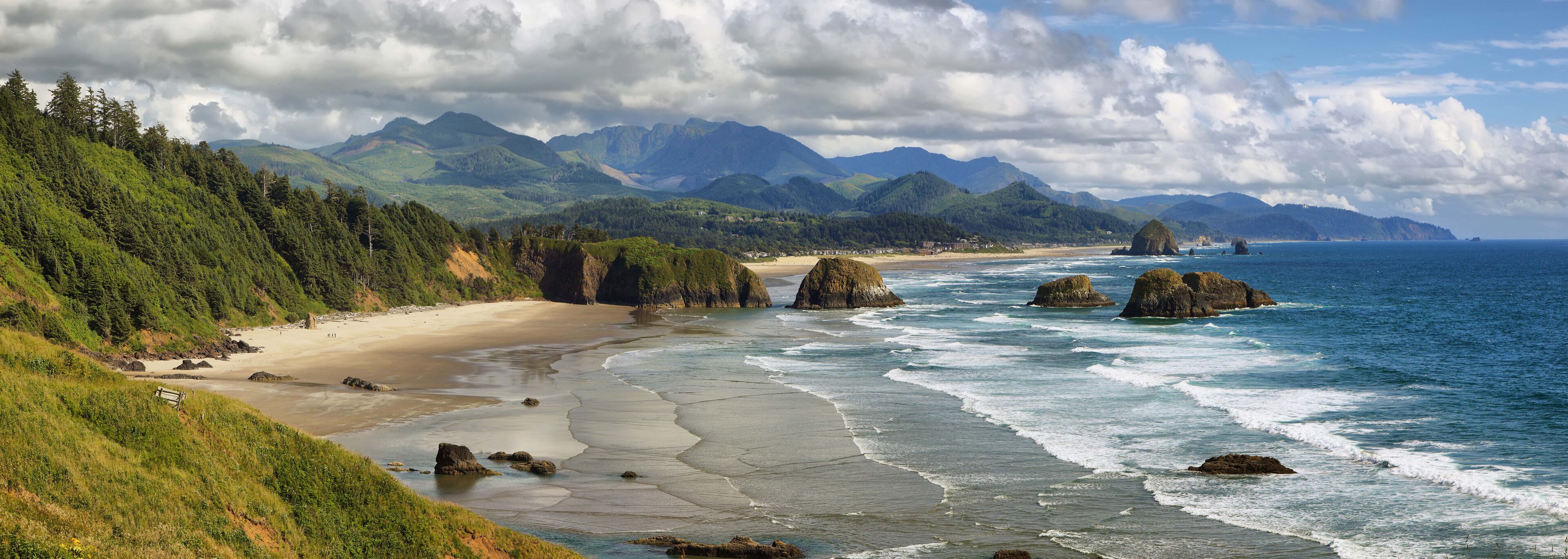 A coastal landscape with a sandy beach, rocky outcrops, and a backdrop of green mountains under a partly cloudy sky.