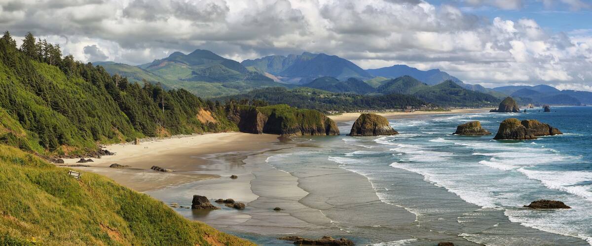 A coastal landscape with a sandy beach, rocky outcrops, and a backdrop of green mountains under a partly cloudy sky.