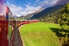 Ein roter Zug fährt durch eine gebirgige Landschaft mit grünen Feldern und blauem Himmel.