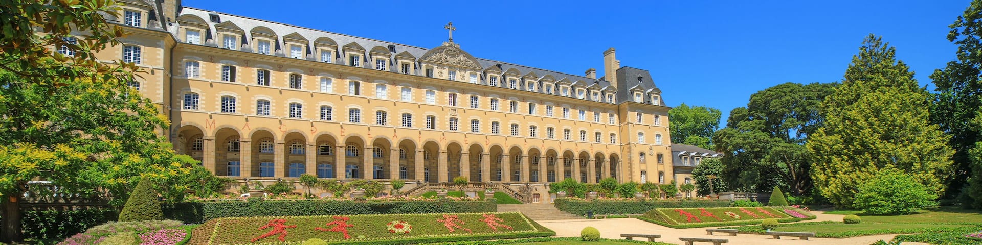 A historic building with a symmetrical facade, surrounded by manicured gardens and a gravel pathway.