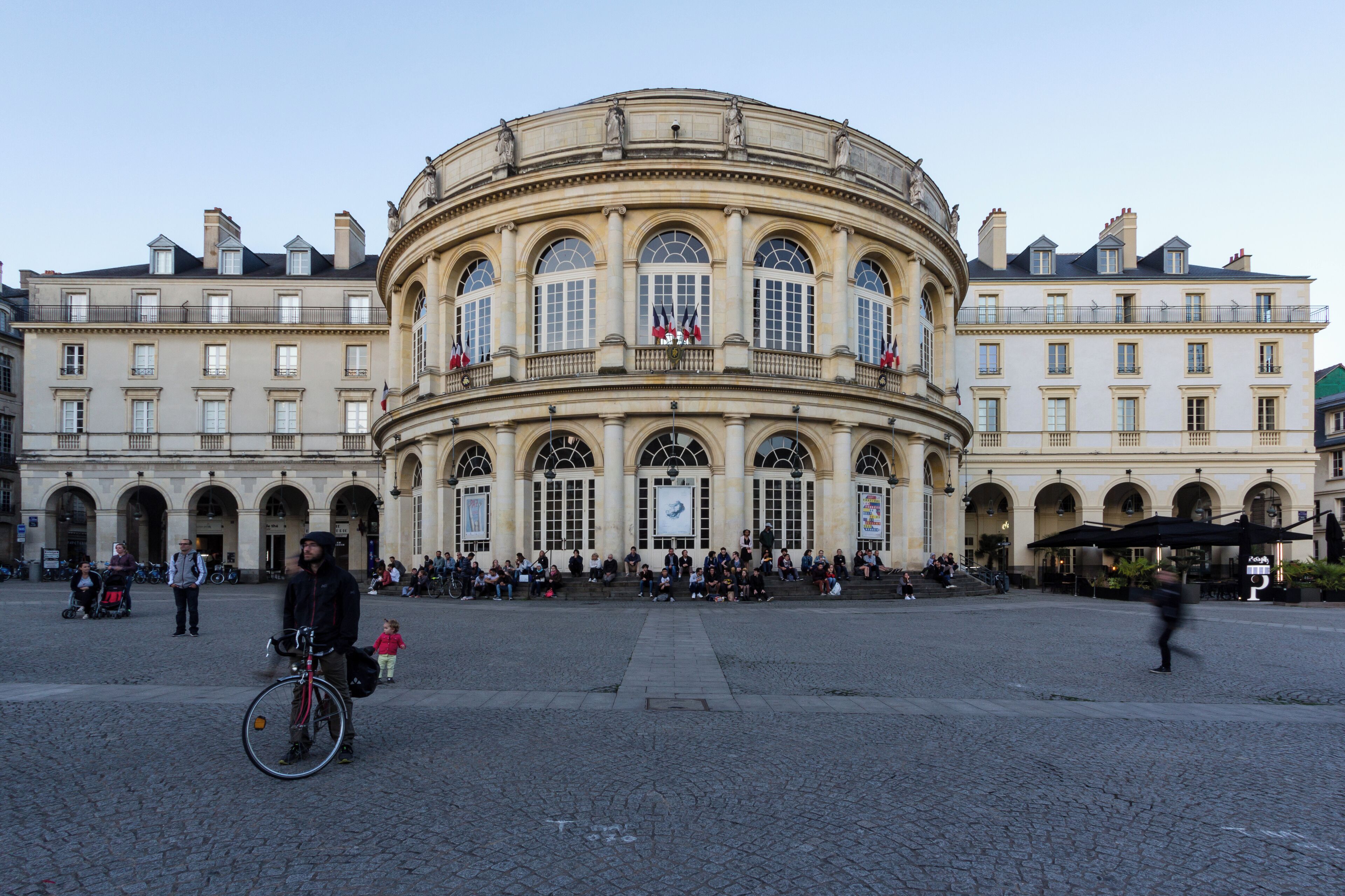 A historic building with arched windows and flags, surrounded by a cobblestone square with people and a bicycle.