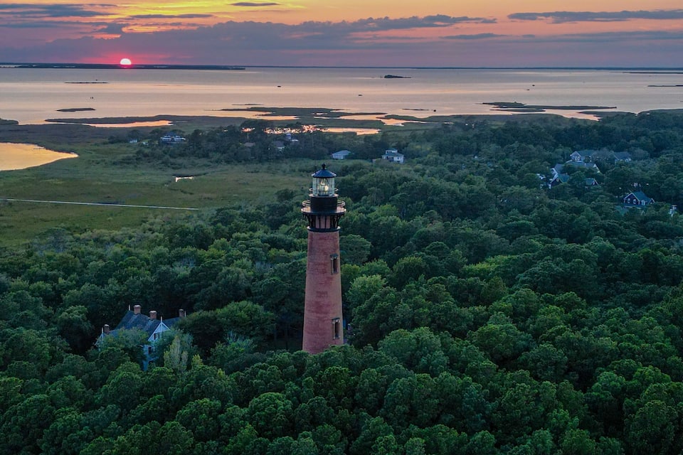 A lighthouse surrounded by dense forest, with a sunset over a calm body of water and distant shoreline.