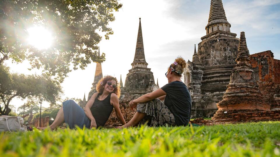 Two women enjoying a sunny afternoon at Ayutthaya Historical Park in Thailand, sitting on grass near ancient temples and stupas, surrounded by nature and cultural heritage