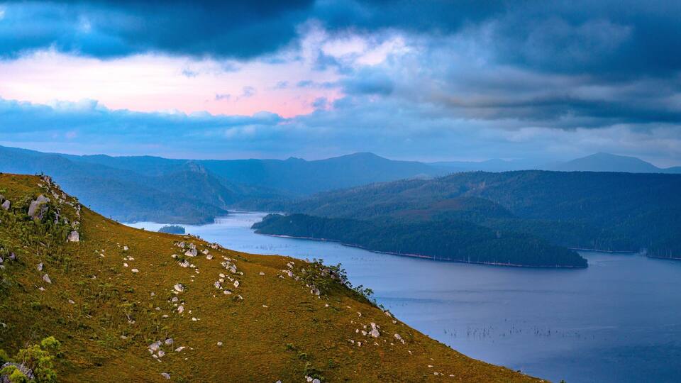 A mountainous landscape with a lake and distant mountains under a cloudy sky.