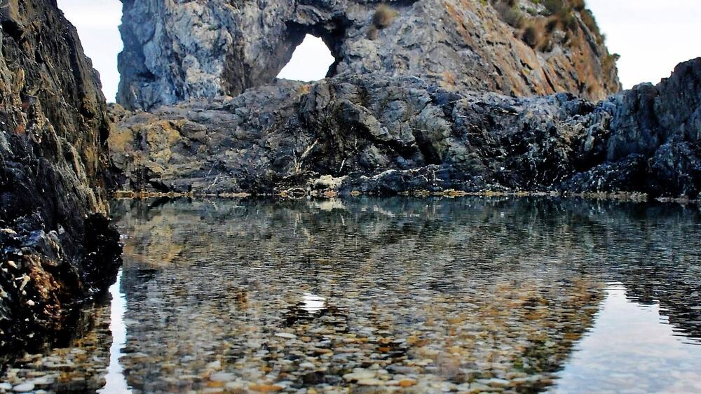 Un paysage côtier rocheux, caractérisé par une arche naturelle imposante et un reflet net dans l’eau.