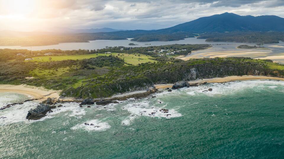 A coastal landscape with a beach, rocky shoreline, and a forested area.