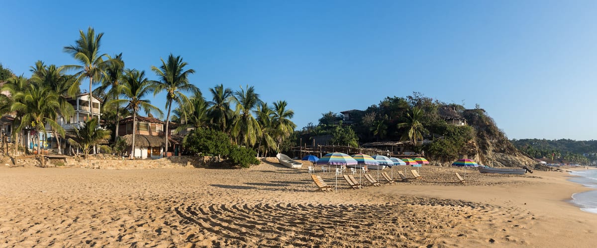A sandy beach with palm trees, beach umbrellas, and a rocky hillside in the background.