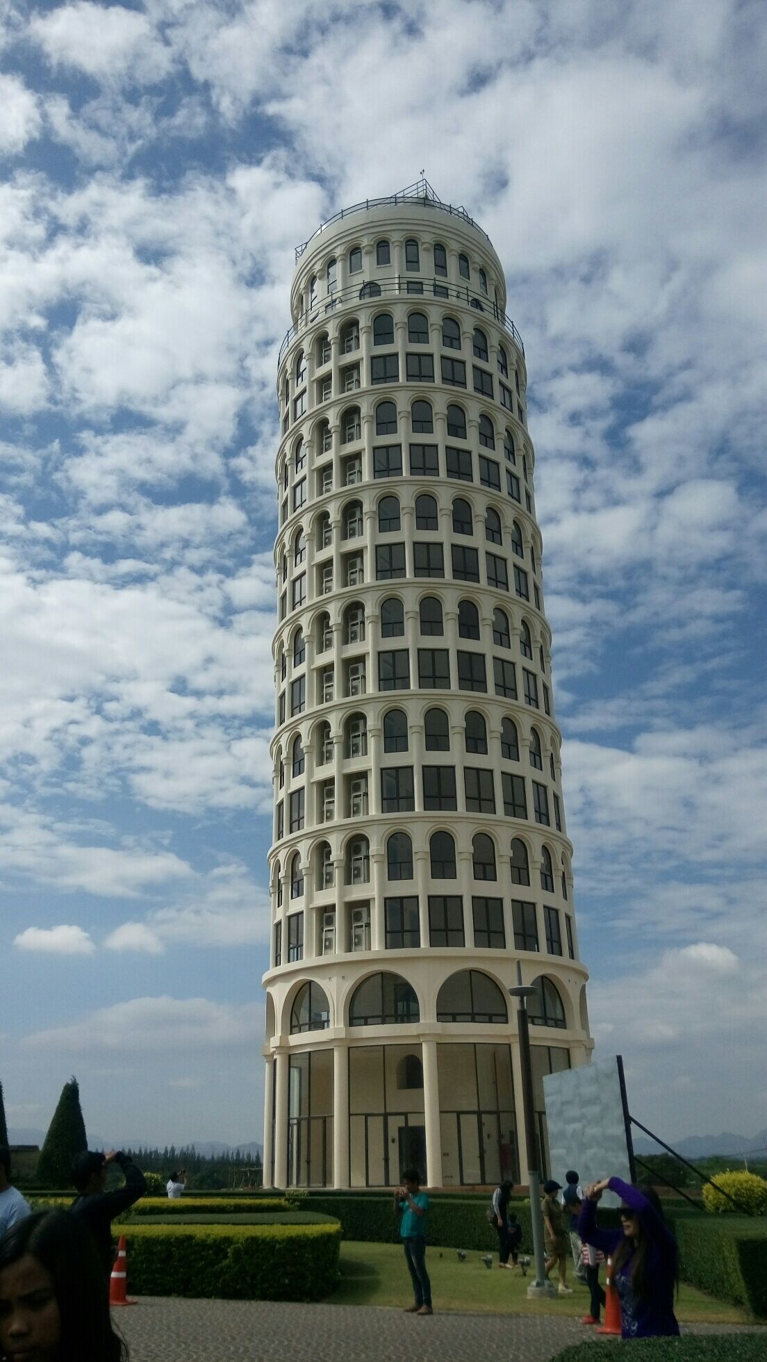 A tall, cylindrical building with arched windows and a flat roof, surrounded by a landscaped area with people and trees.