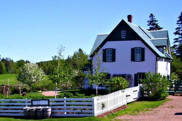 A white house with a green roof, surrounded by a white picket fence and lush greenery.
