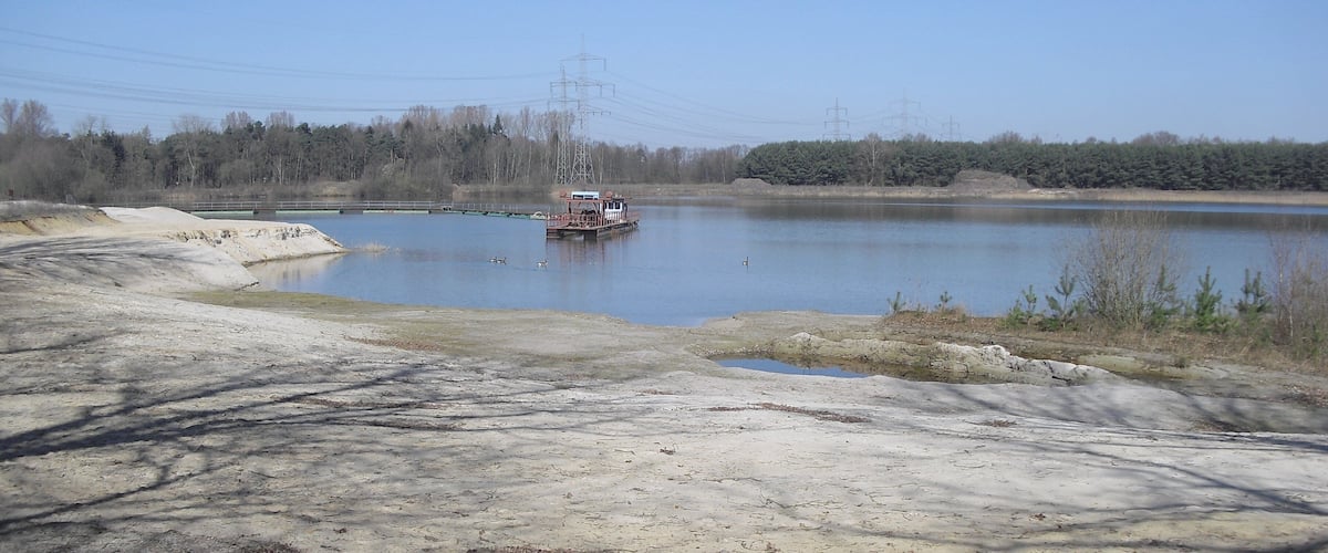 A boat on a calm lake with a sandy shore and power lines in the background.