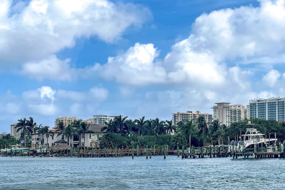 A waterfront cityscape with palm trees, a marina, and high-rise buildings under a partly cloudy sky.