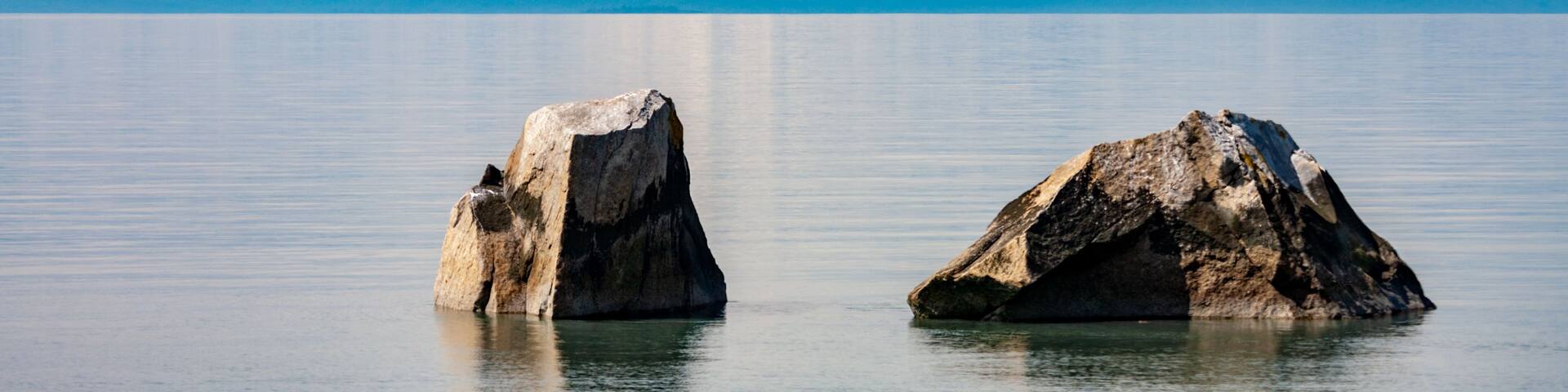 Montaña nevada con afloramientos rocosos en primer plano y una masa de agua tranquila que refleja la escena.