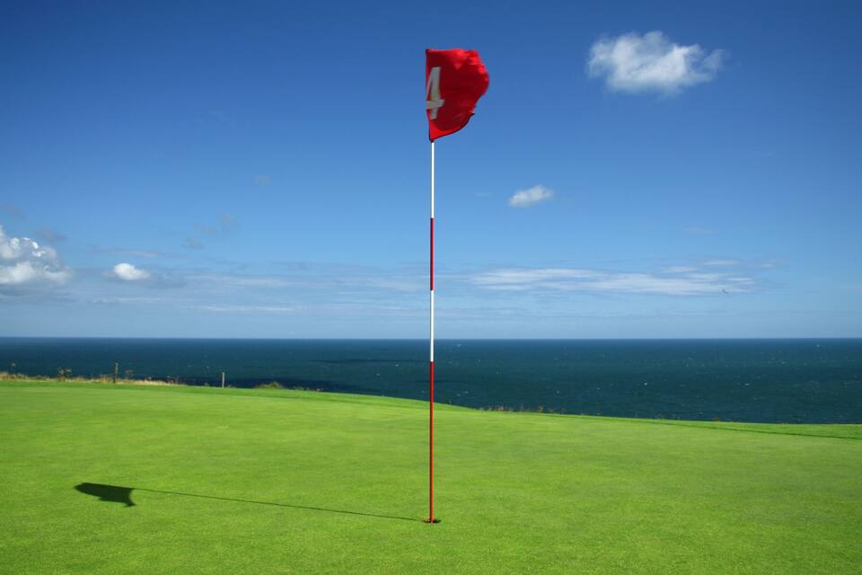 A golf course with a flag on a pole, ocean in the background, and a clear sky.