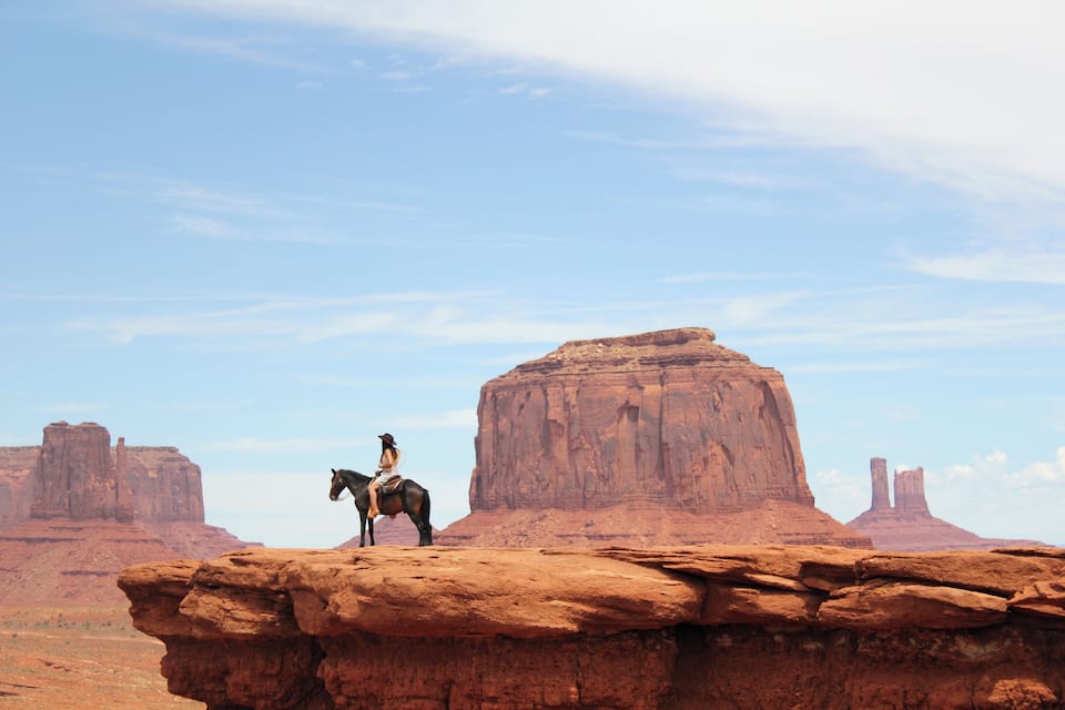 A cowboy on a horse atop a rock formation in a desert landscape with multiple buttes in the background.