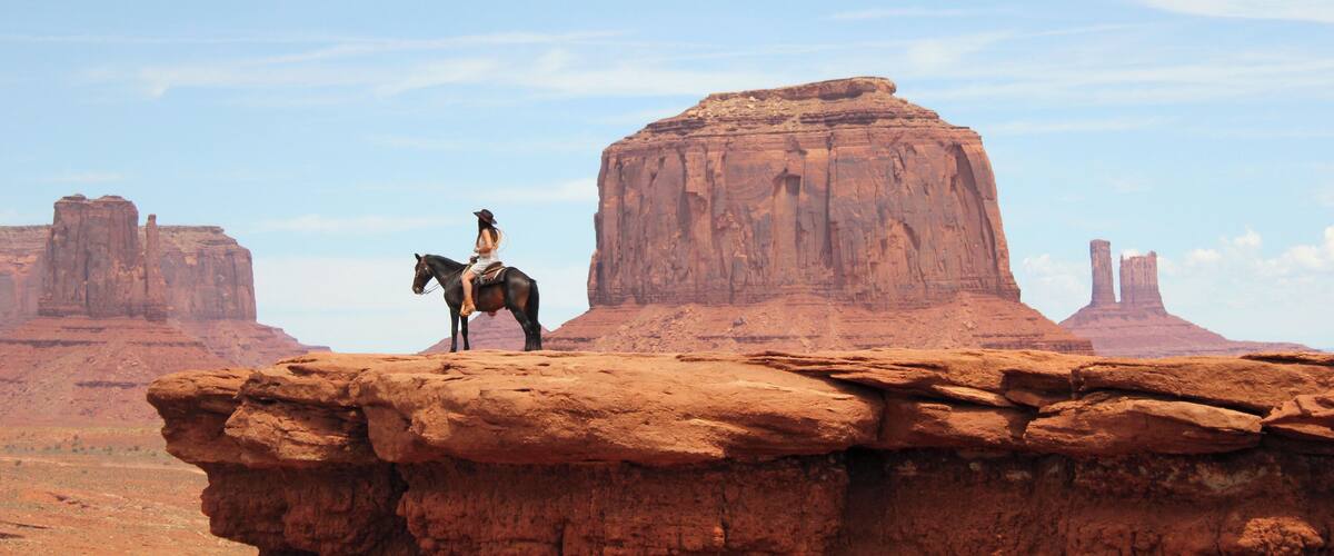 A cowboy on a horse atop a rock formation in a desert landscape with multiple buttes in the background.