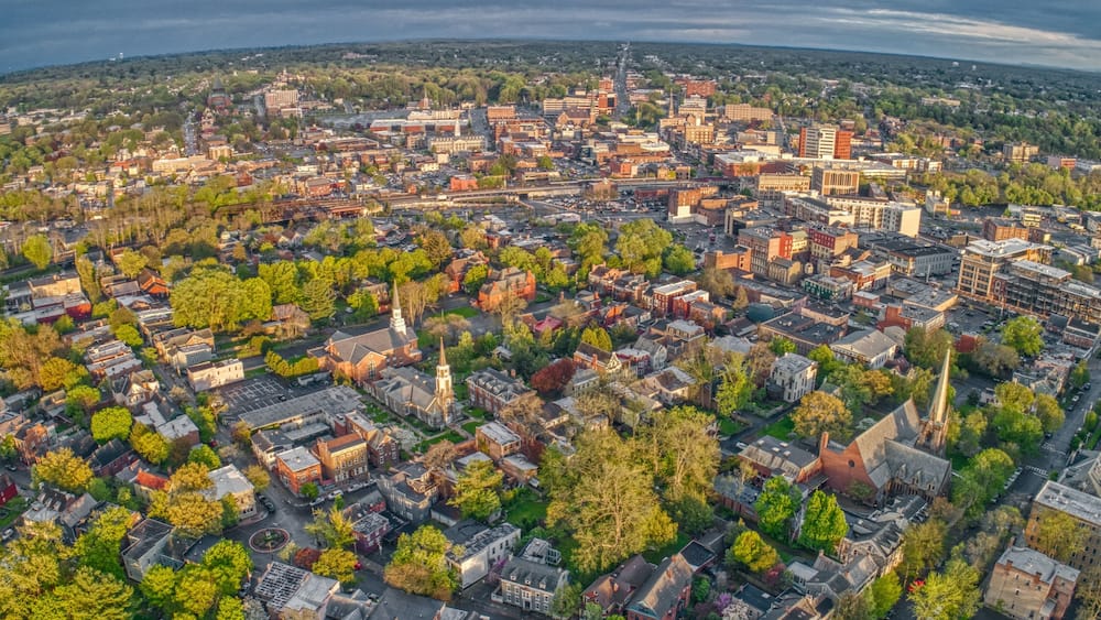 An aerial view of a city with a mix of historic and modern buildings, surrounded by green spaces and trees.