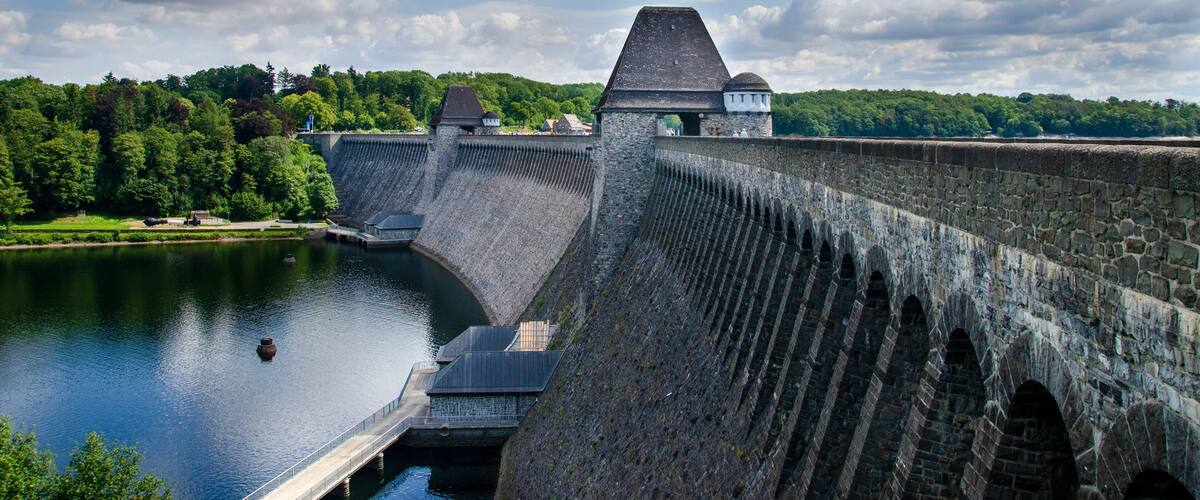 A large dam with a sloped top and multiple arches, surrounded by a forested area and a body of water.