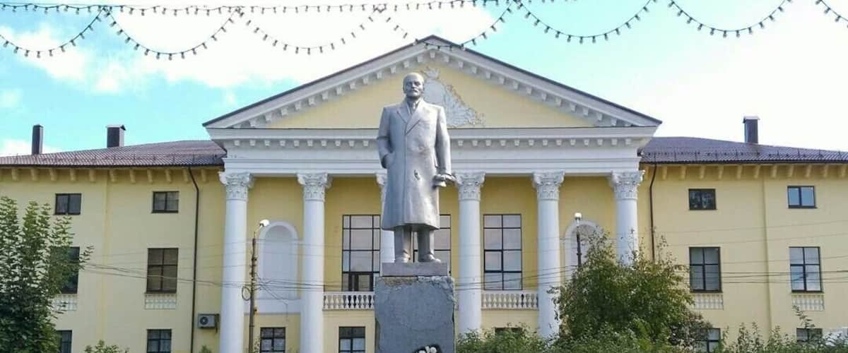 A statue in front of a yellow building with columns, surrounded by flowers and pigeons.