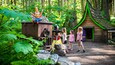 A forest scene with a wooden structure, a large doll, and a green-roofed building.