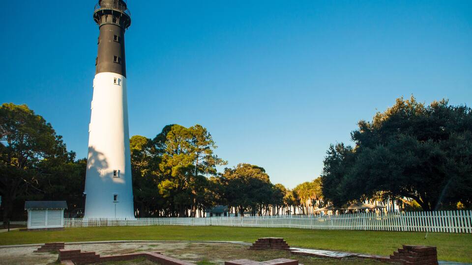 A lighthouse with a white and black tower, surrounded by a white picket fence, trees, and a grassy area with brick steps.