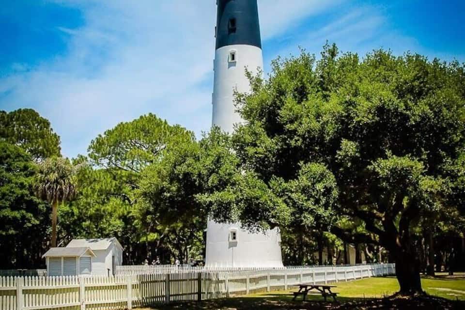 A lighthouse with a white and black tower, surrounded by trees and a white fence.