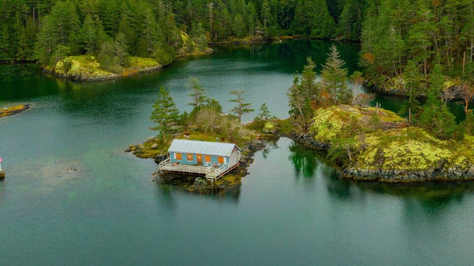 A small house on a rocky island surrounded by a calm lake and dense forest.