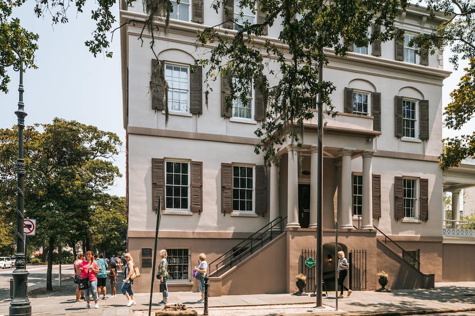 A historic building with brown shutters and columns, surrounded by trees and pedestrians.