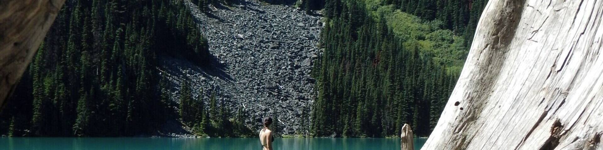 A person standing on a log over a turquoise lake with mountains in the background.