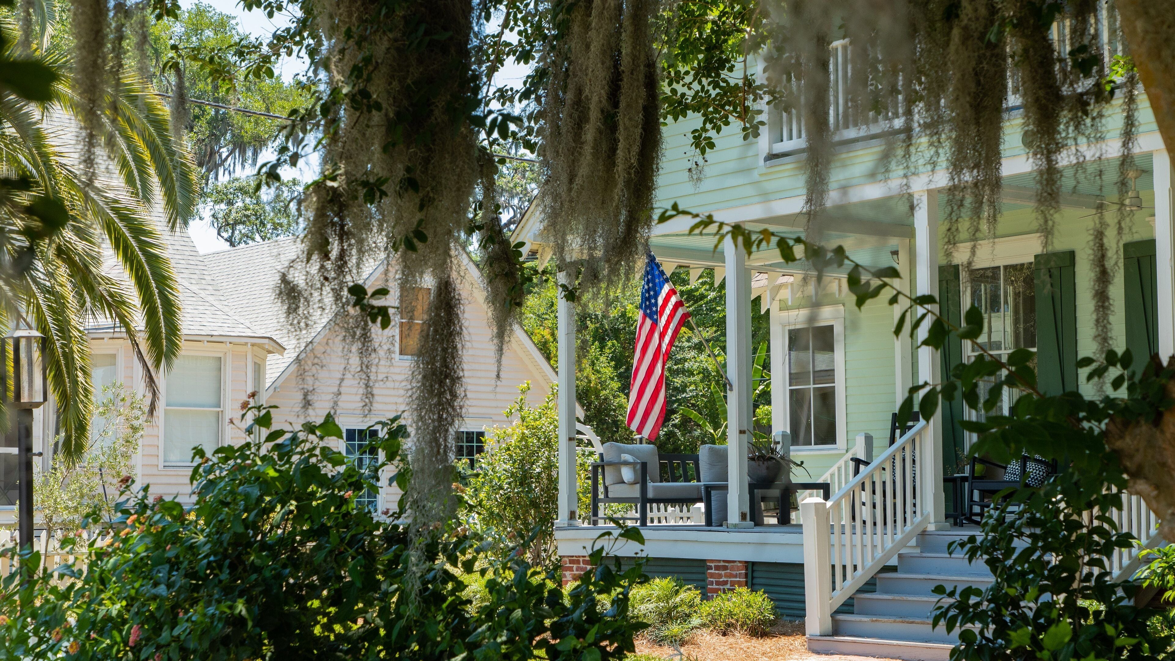 A porch with an American flag, surrounded by lush greenery and a house with a green roof.