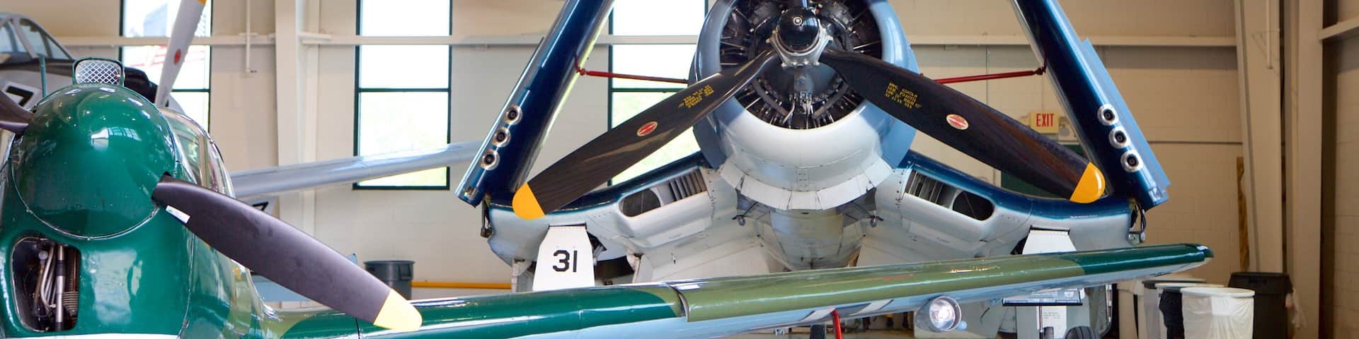 Un avion d’époque, avec un fuselage avant vert et des ailes bleues, est exposé dans un hangar, aux côtés d’autres avions.