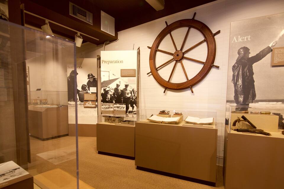 Una mostra museale con manufatti storici, tra cui un grande timone di nave, documenti e una statua.